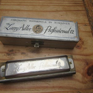 Könnte beinhalten: Ein chromatisches Vintage-Mundharmonika von M. Hohner, die "Larry Adler Professional 12", mit Originalkoffer. Die Mundharmonika ist silberfarben, und der Koffer ist hellgrau mit dem Text "Made in Germany".