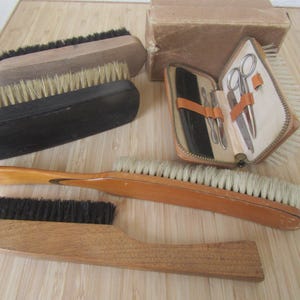 May include: An assortment of grooming items, featuring wooden brushes with bristles and a small leather case with manicure tools. The brushes vary in shape and size, some with black bristles and others with light-coloured bristles. The case is open, revealing the tools.