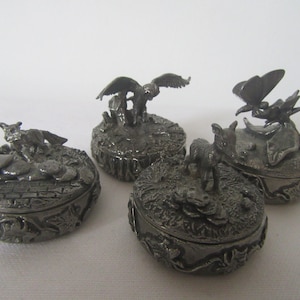May include: Four pewter trinket boxes with animal figurines on top. The boxes are oval shaped and have a detailed design. The animals include a fox, a bird, a butterfly, and a pig.