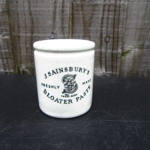 Könnte beinhalten: Ein weißer Keramiktopf mit Deckel, mit dem schwarzen Text "J. Sainsbury's Freshly Bloater Paste". Der Topf hat eine zylindrische Form und ist wahrscheinlich ein antiker Lebensmittelbehälter.