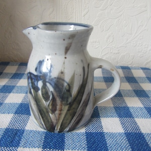 Op de afbeelding: Een keramische kan met een witte gespikkelde glazuur, met een handgeschilderd ontwerp van blauwe irissen en groene bladeren. De kan heeft een handvat en een tuit en staat op een blauw-wit geruit tafelkleed.