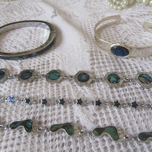 Puede incluir: Una colección de joyas que incluye una pulsera de plata con incrustaciones de abulón, una pulsera de plata con una piedra azul y varias pulseras de cadena de plata con detalles de abulón y forma de estrella. Las joyas se exhiben sobre una superficie de encaje blanco.
