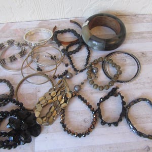 Op de afbeelding: Een verzameling armbanden en bangles in verschillende stijlen en materialen, waaronder zwart, bruin, goud, zilver en grijs. Sommige armbanden zijn versierd met kralen, terwijl andere van metaal of hout zijn gemaakt.