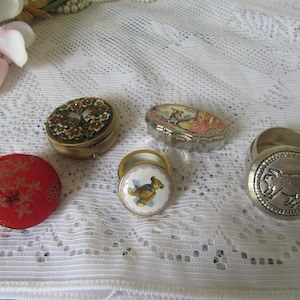 May include: An assortment of small, decorative trinket boxes and a ring holder. The boxes include a red fabric box, a gold-toned box with floral details, and a silver-toned box with a dog illustration. The ring holder has a pig design.