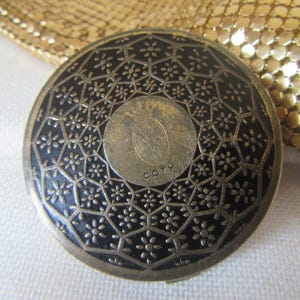 May include: A vintage Coty compact with a black and silver floral design. The round compact features a central silver disc with the Coty logo and text. The background is a gold mesh purse.