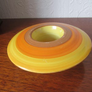 May include: A yellow, orange, and brown ceramic candle holder with a circular base and a wide, shallow bowl. The holder has a layered, concentric design.