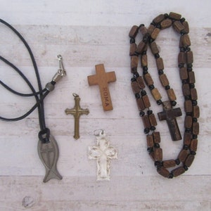 May include: A collection of five religious pendants, including a wooden cross with the word "ROMA" inscribed on it, a silver fish pendant, a wooden rosary with a wooden cross, and two silver crosses.
