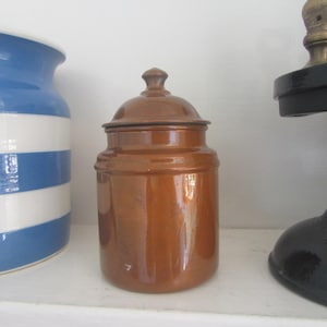 Puede incluir: Un tarro de cerámica marrón con tapa. El tarro es cilíndrico con una base redondeada y un pequeño pomo en la parte superior. A la izquierda se ve parcialmente un tarro a rayas azules y blancas.