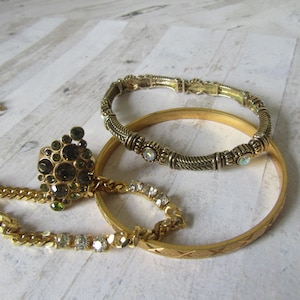 May include: A collection of gold-toned jewelry including a ring with dark green stones, a textured bracelet, a bangle, and a chain necklace with clear stones. The jewelry is arranged on a light-colored, patterned surface.