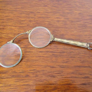 Op de afbeelding: Een paar antieke zilverkleurige vouwbrillen met ronde glazen. De bril heeft een decoratief scharnier en een sluiting.