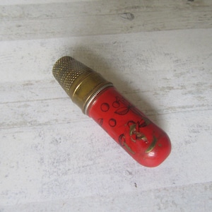 Puede incluir: Un portadedal rojo vintage con tapa y detalles de latón. El soporte cilíndrico presenta un diseño pintado de hojas y bayas doradas. Un accesorio de costura único.