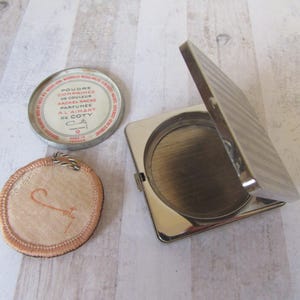 Könnte beinhalten: Vintage Silber-Kompakt mit einem weißen und roten Etikett mit der Aufschrift "Poudre Compressee en Couleur Rachel Rache Parfumée Al Aimant de Coty". Das Kompakt ist geöffnet und der Spiegel ist sichtbar. Es gibt einen runden, weißen Puderquast mit rosa Einfassung.