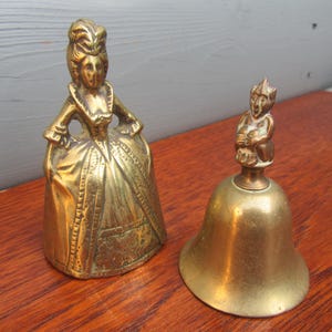 May include: Two brass bells. One bell is shaped like a woman in a dress, the other is a bell with a small figure on top. Both bells are gold in color and sit on a wooden surface.