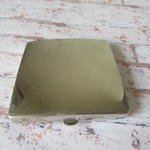 Op de afbeelding: Een vintage, vierkante, zilverkleurige compact case. De compact heeft een licht afgeronde bovenkant en een kleine sluiting aan de voorkant. Het oppervlak vertoont lichte slijtage, wat op leeftijd duidt. De achtergrond is een rustiek, verweerd houten oppervlak.