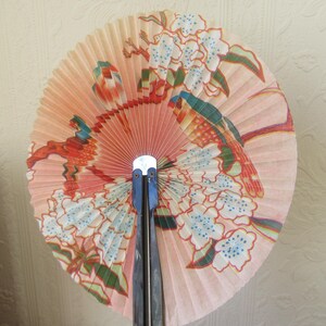 May include: A pink and white folding fan with a floral design and two birds. The fan is made of paper and has a black wooden handle.