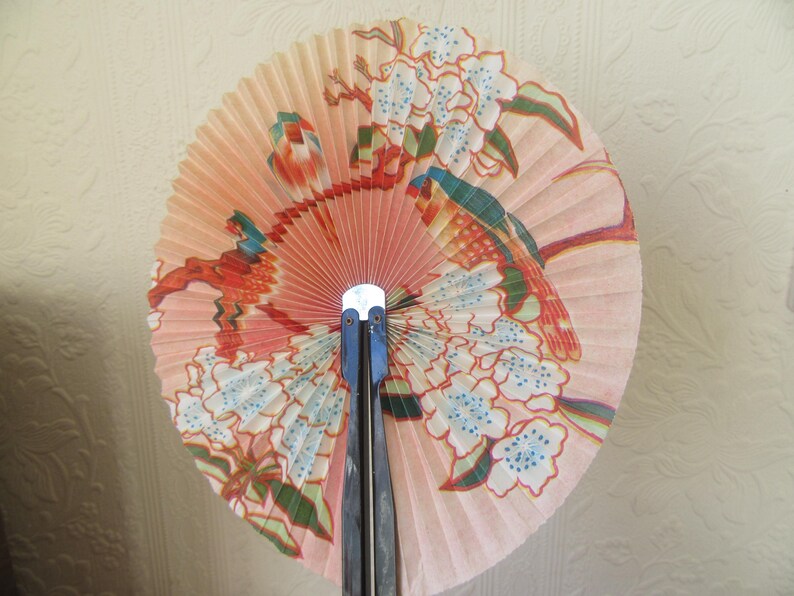 May include: A pink and white folding fan with a floral design and two birds. The fan is made of paper and has a black wooden handle.