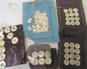 Op de afbeelding: Een assortiment vintage knopen en versieringen op verouderde kaarten. De knopen zijn parelmoer, en de kaarten zijn in verschillende kleuren, waaronder blauw en zwart. Sommige kaarten zijn gemarkeerd met "The Windsor Card."