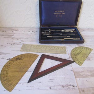 May include: A vintage wooden box containing a set of drafting tools, including a compass, protractor, and ruler. The box is labelled "The Hathbone Technical Case No. 92."