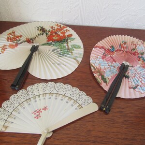 May include: Three decorative folding fans with floral designs. The fans are made of paper and have black and pink handles.