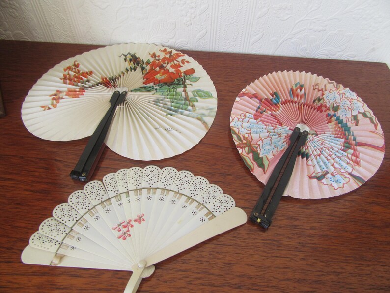 May include: Three decorative folding fans with floral designs. The fans are made of paper and have black and pink handles.