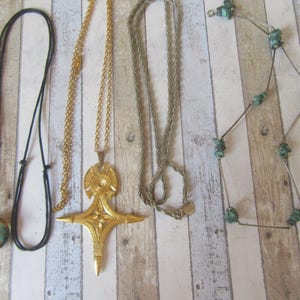 Puede incluir: Una colección de collares con varios diseños. Incluye un collar de cordón negro con un colgante turquesa y dorado, una cadena dorada con un colgante de cruz, una cadena plateada y un collar plateado con piedras turquesas.