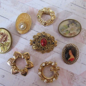Könnte beinhalten: Sortiment von acht Broschen im Vintage-Look in Goldtönen mit verschiedenen Designs, darunter florale, Porträt- und geometrische Muster. Einige Broschen sind mit Kunstperlen und Strasssteinen verziert.