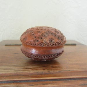 Peut inclure: Une petite boîte ronde en bois avec des motifs sculptés complexes. La boîte est brune et a un couvercle qui s'ajuste parfaitement sur le dessus.