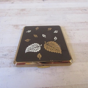 May include: A gold-colored compact with a brown background and a design of white and gold leaves.