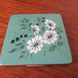 Op de afbeelding: Een vintage compact met een bloemenontwerp. De compact is gemaakt van metaal en heeft een lichtblauwe achtergrond met witte bloemen en groene bladeren.