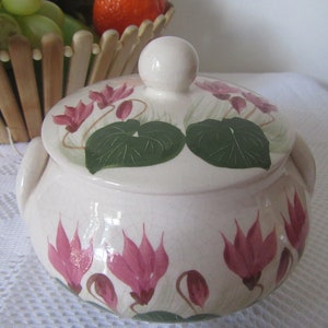 May include: A white ceramic covered dish with a floral design. The dish is decorated with pink cyclamen flowers and green leaves. The lid has a knob on top.