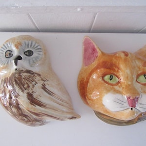 Könnte beinhalten: Zwei keramische Wandplatten mit einem handbemalten Eulen- und einem Katzenmotiv. Die Eule hat braune und weiße Federn mit einem grauen Gesicht. Die Katze hat orangefarbenes Fell, grüne Augen, rosa Ohren und eine weiße Schnauze.