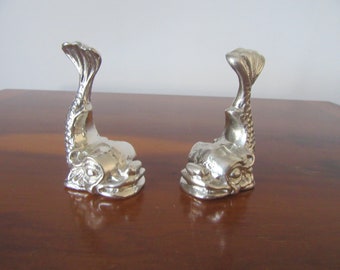 May include: Two silver fish-shaped decorative table accents. The fish are facing opposite directions with their tails raised.