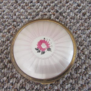 Può includere: Un compatto rotondo vintage con una cornice in metallo dorato. Il coperchio presenta un design rosa e bianco con un motivo floreale centrale. Il compatto viene probabilmente utilizzato per contenere la cipria.