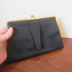 May include: A black satin clutch with a gold-toned frame. The rectangular bag has a simple design with a flap closure and a small handle. The clutch is held in a hand, suggesting its size and portability. A vintage accessory.