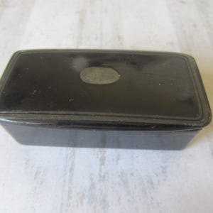 May include: A rectangular black box with a slightly raised lid. A small, oval-shaped, silver-colored accent is centered on the lid. The box has a simple, classic design, and appears to be made of a solid material.