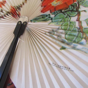 May include: A white folding fan with a floral design featuring red flowers and green leaves. The fan has a black handle and the text "Made in the People's Republic of China" is printed on the fan.
