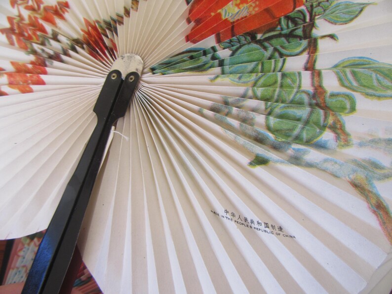 May include: A white folding fan with a floral design featuring red flowers and green leaves. The fan has a black handle and the text "Made in the People's Republic of China" is printed on the fan.
