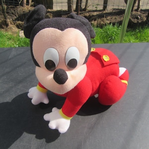 May include: A soft toy of Mickey Mouse in a crawling pose. The toy is dressed in a red outfit with yellow trim, black ears, a black nose, and large eyes. It is positioned on a dark surface.