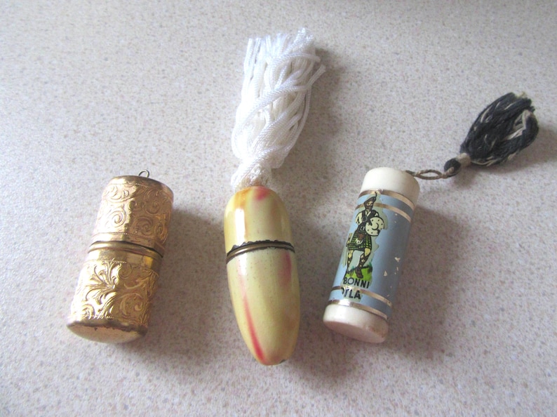 May include: Three vintage thimble cases. One is gold with an ornate design, one is white with a yellow and red design, and one is blue with a white and red design and the text "BONNI VILLA".
