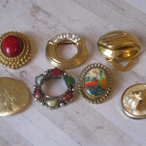 May include: A collection of seven vintage gold-tone brooches with various designs. The brooches feature a red stone, a floral pattern, a horse head, and a circle of colorful stones.