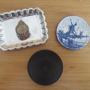 May include: A rectangular silver-toned scalloped tray containing a commemorative metal badge featuring King George and Queen Mary, a circular ceramic coaster with a blue and white windmill scene, and a plain black circular disc object.
