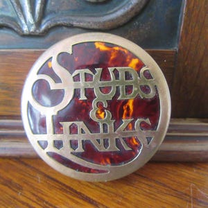 May include: A round, brass-colored pin with a red and orange tortoiseshell design. The pin features the words "Stubbs & Hanks" in raised lettering.