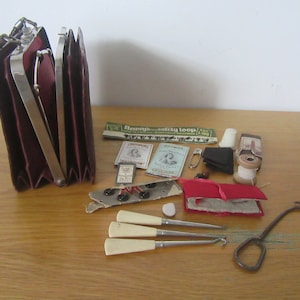 May include: A vintage red leather sewing kit with a hinged clasp and a metal handle. The kit is open and contains various sewing supplies, including needles, thread, buttons, a thimble, and a small pair of scissors.