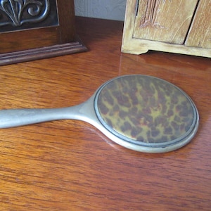 Op de afbeelding: Een zilverkleurige handspiegel met een glazen oppervlak met een schildpadpatroon. De spiegel heeft een eenvoudig, elegant ontwerp.