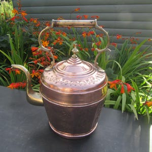 May include: A copper tea kettle with a polished finish, featuring an ornate brass handle and a curved brass spout. The lid has a pointed finial, and the body is decorated with embossed floral patterns around the upper rim and base section.