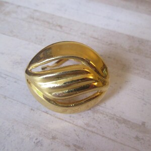 May include: A gold-toned metal earring with a wavy, abstract design. The earring is circular and has a cutout in the center.