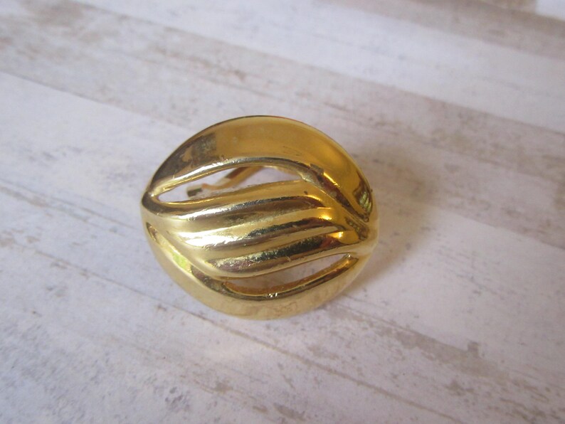 May include: A gold-toned metal earring with a wavy, abstract design. The earring is circular and has a cutout in the center.