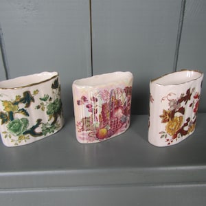 May include: Three white ceramic vases with a gold rim. Each vase features a different floral design in shades of green, yellow, red, and brown. The vases are arranged on a gray surface.