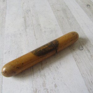 Puede incluir: Objeto cilíndrico de madera con acabado marrón claro. El objeto tiene una banda oscura alrededor del centro y un área ligeramente más oscura con una imagen. Está sobre una superficie de madera clara.