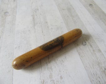 May include: A cylindrical wooden object with a light brown finish. The object has a dark band around the middle and a slightly darker area with an image on it. The object is lying on a light-colored wooden surface.